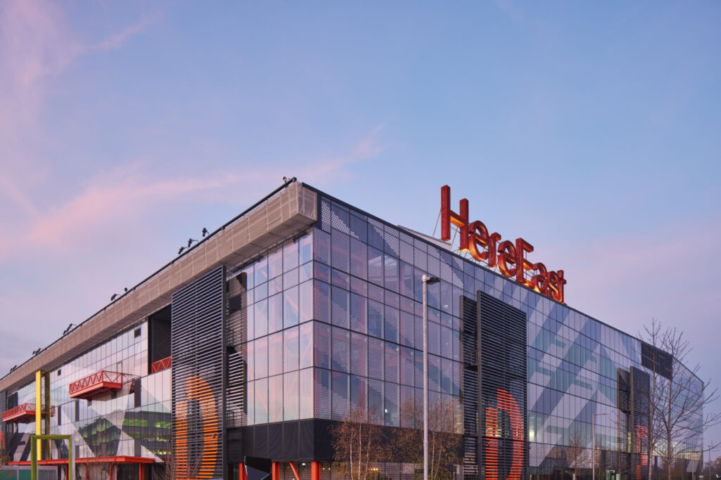 A modern office building featuring lots of external glazing with a large red Here East sign on top