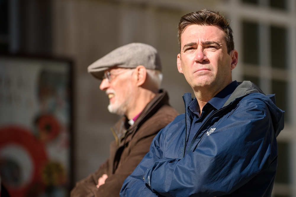 Andy Burnham wears a blue coat and looks off into the distance. He stands alongside an older man wearing a flat cap