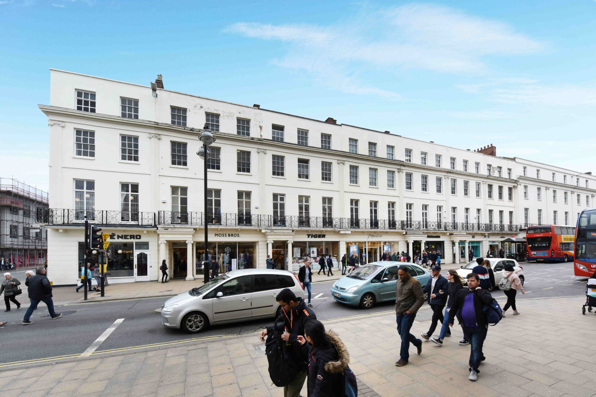 A parade of shops housed in a terraced building with a white facade with cars and pedestrians in the foreground