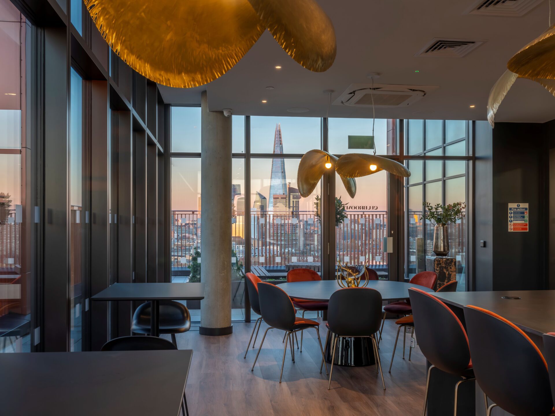 Interior of a building featuring columns and windows, hanging lights, a series of desks and chairs and a view of office towers in the distance