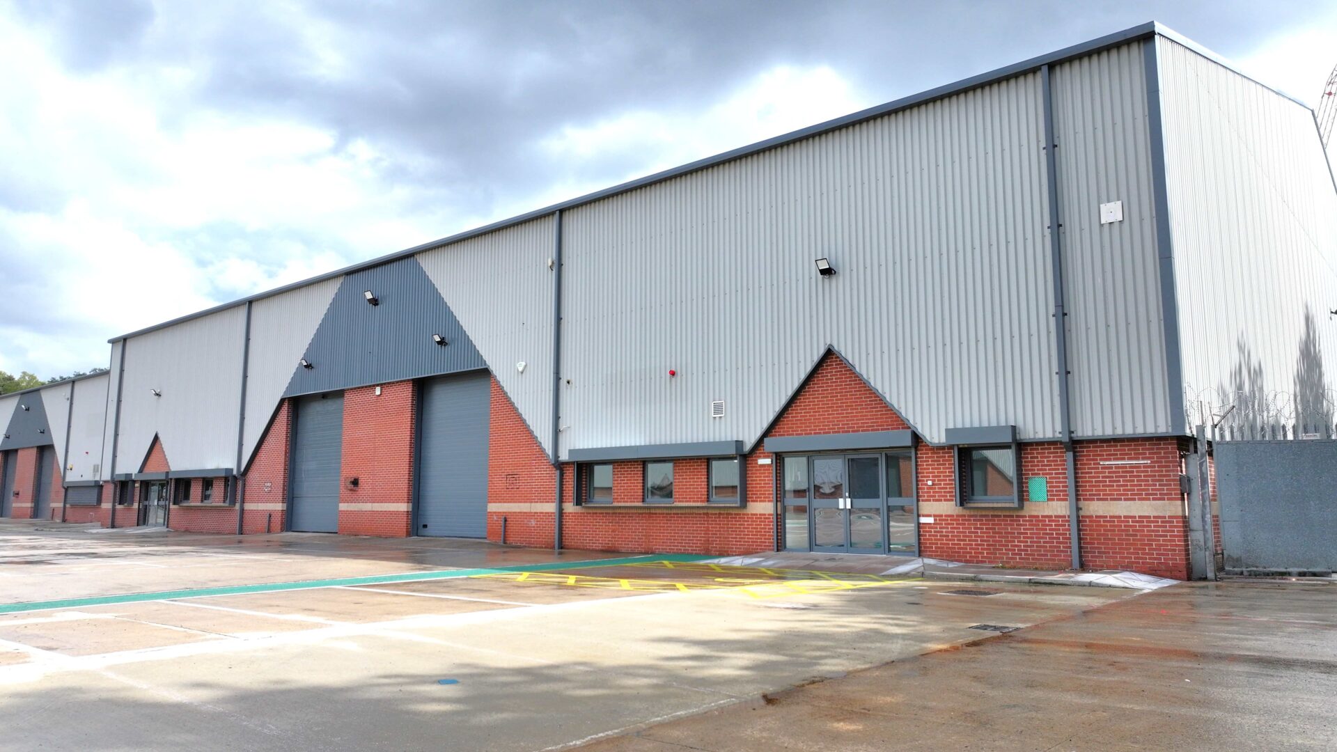 A row of brick and steel cladded industrial buildings with car parking and yard space in the foreground