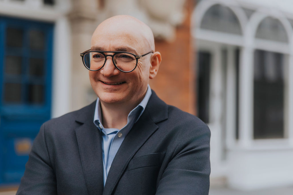 Ilan Goldman is a white, bald man wearing black rimmed glasses, a dark blue suit and a collared light blue shirt