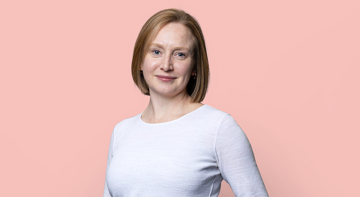 Liz Turner has a bob and is stood against a peach background. She is wearing a light top and dark trousers