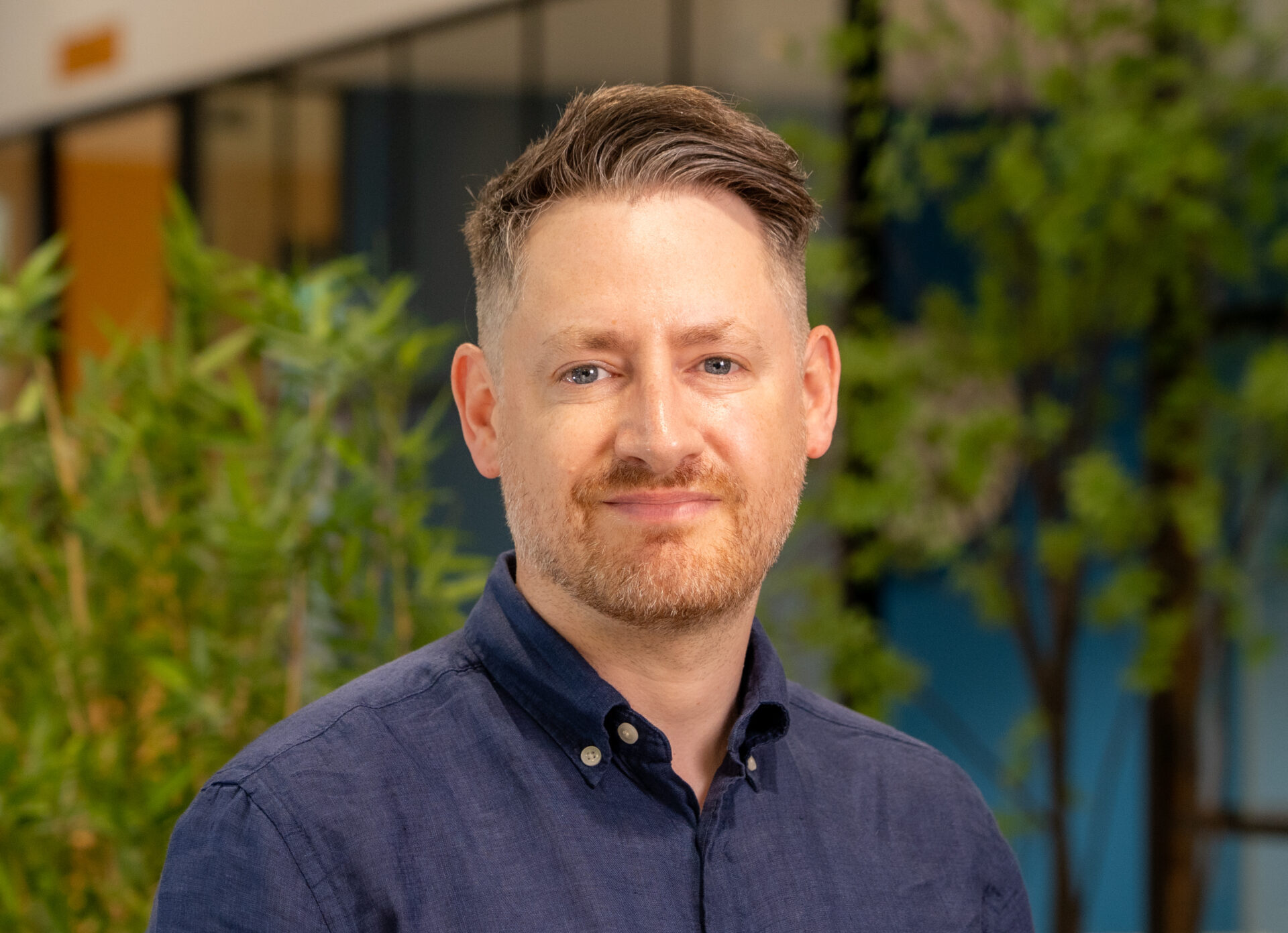 A while male wearing a dark blue collared shirt with trees in the background