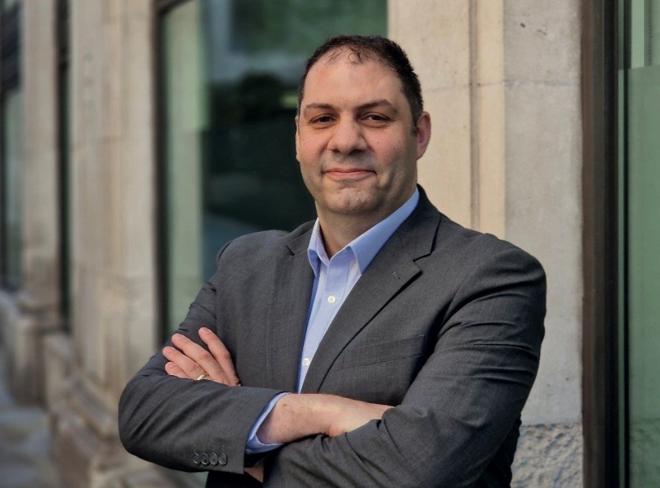 A man wearing a grey suit and a light blue shirt with his arms folded