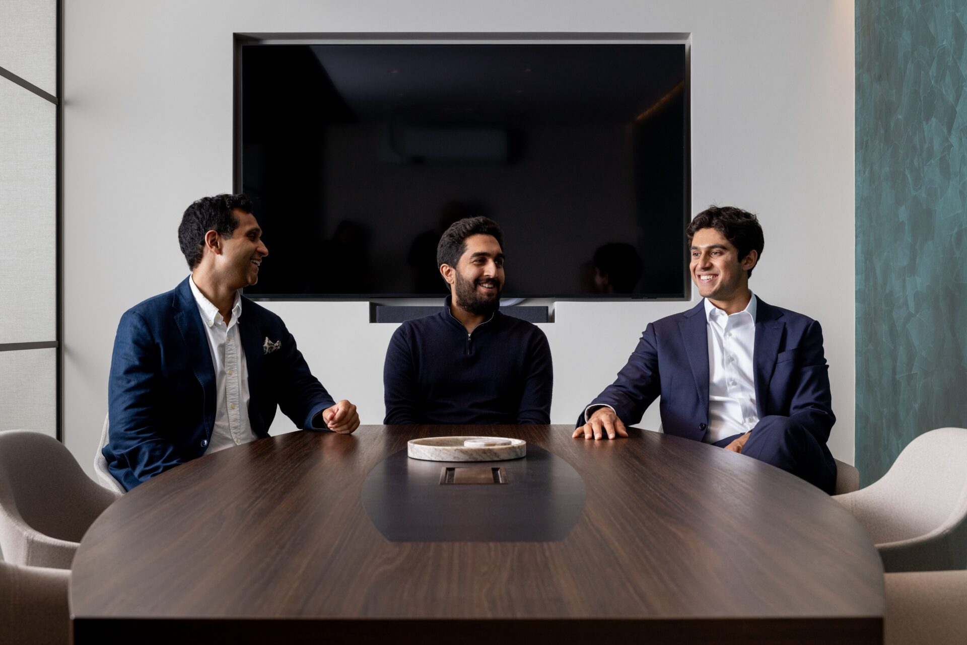 Three smartly dressed men sat around a table with a large TV screen behind them