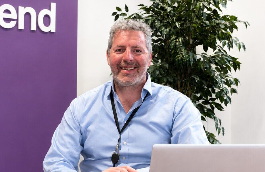 Picture of a smiling man wearing a blue shirt sat behind a table with a computer