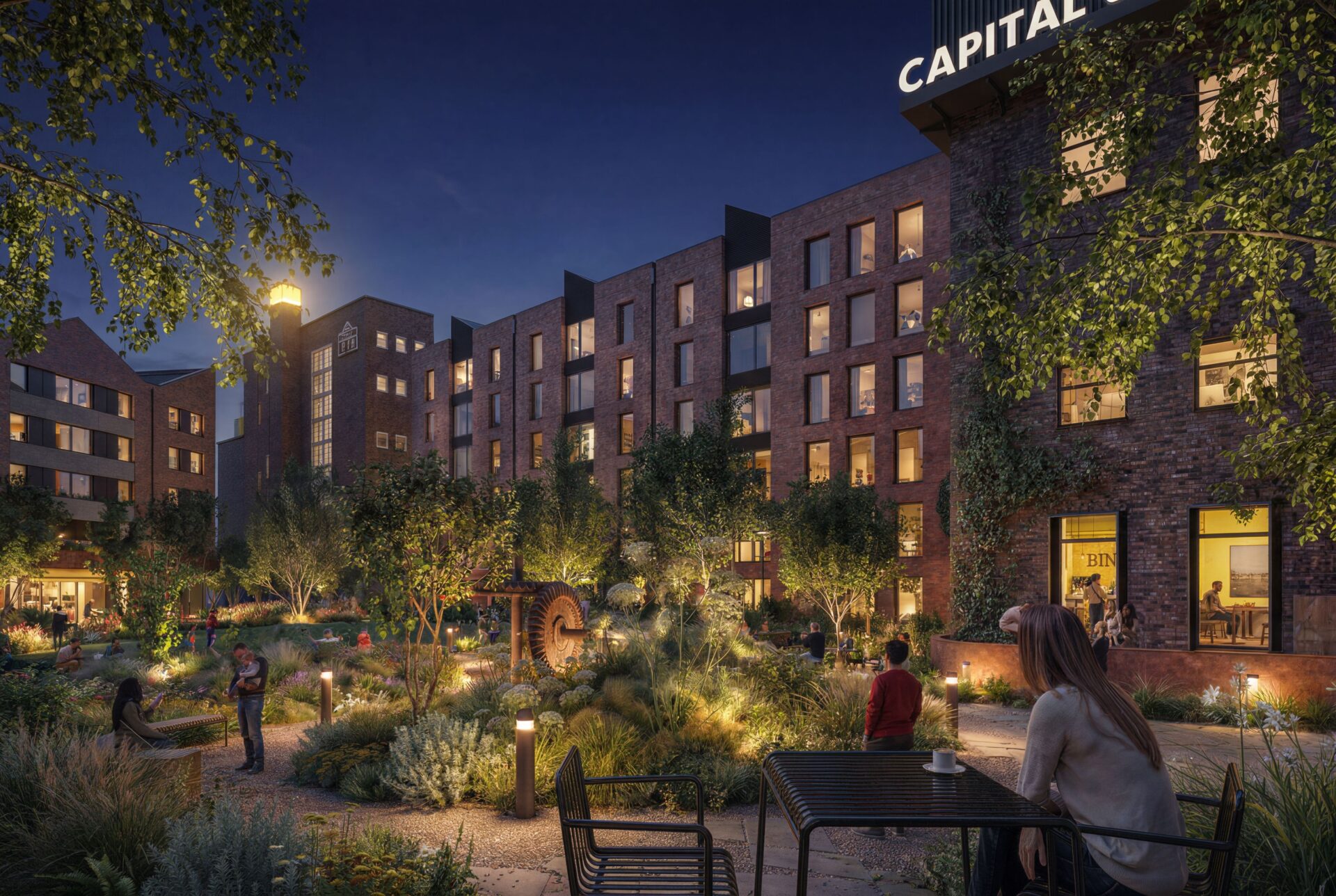 Image of a series of brick built buildings at night with a garden and seating in the foreground