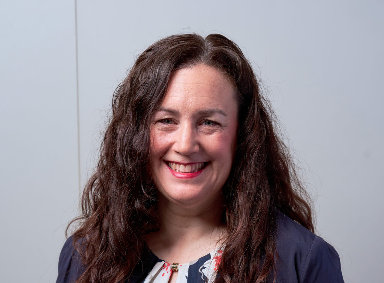 A smiling woman with dark long curly hair wearing a blue jacket and blue dress with a flower print