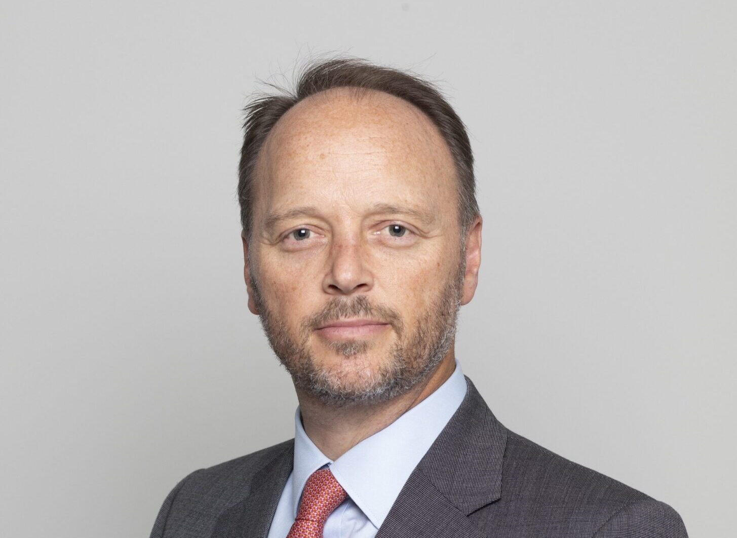 A white male wearing a grey suit jacket, a light shirt and a red tie