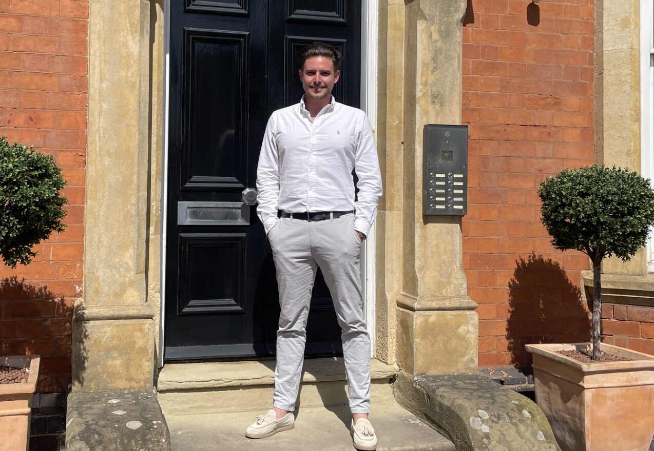 Picture of a white male wearing a white collared shirt and light coloured trousers stood outside the doorway of a red brick building