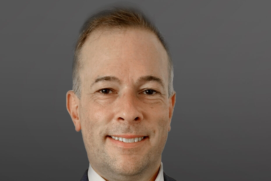A smiling man with short hair wearing a dark suit jacket and a white shirt