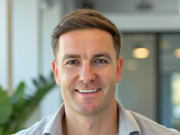 A smiling man with short brown hair wearing a collared grey shirt