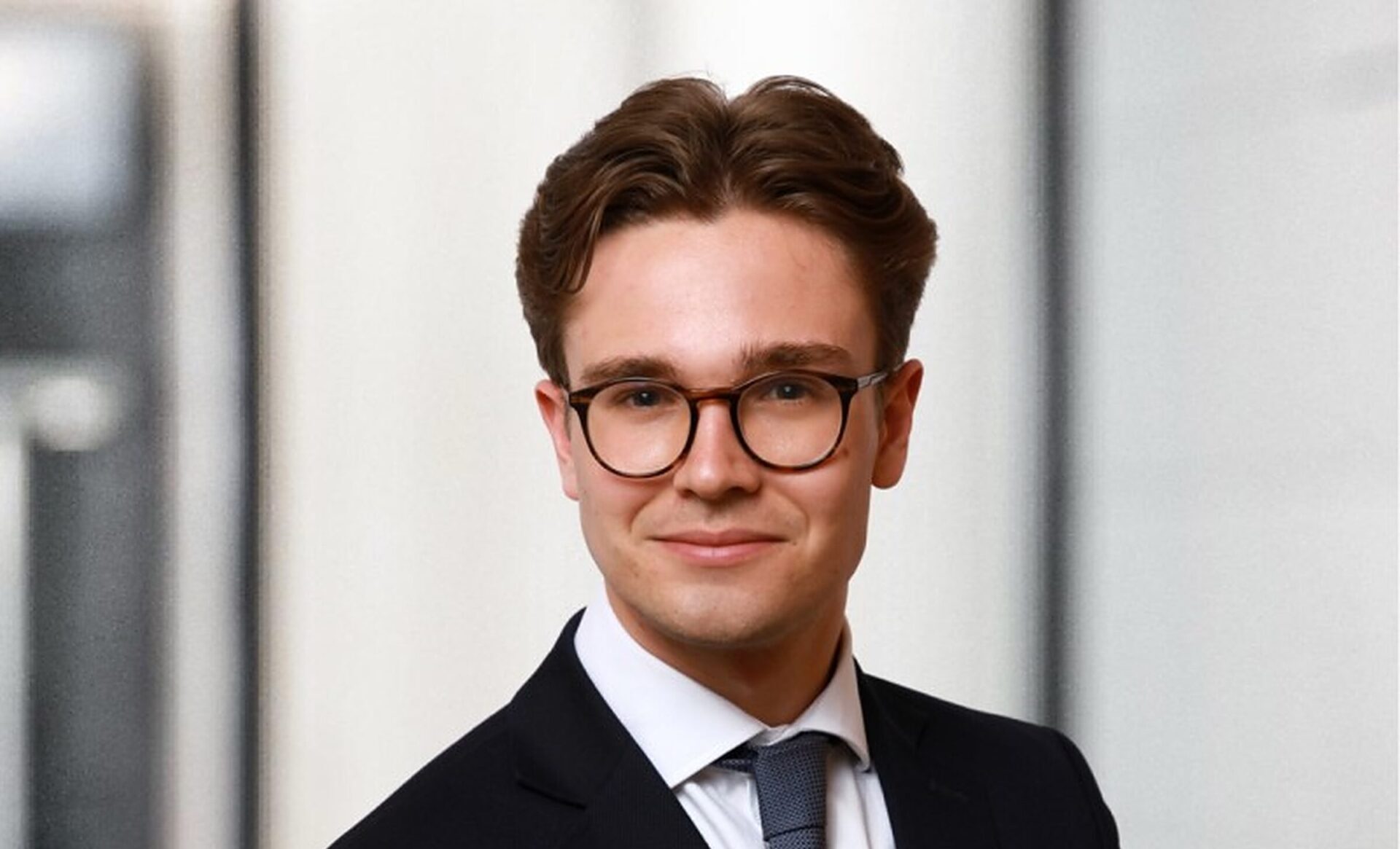 A man with dark brown hair wearing glasses and a dark suit with a white shirt and tie