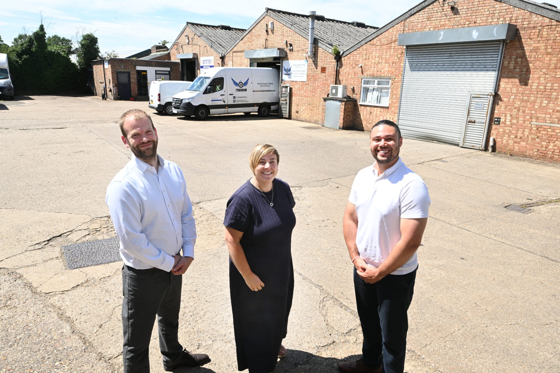 Two men and a woman stood in a yard in front of a warehouse building