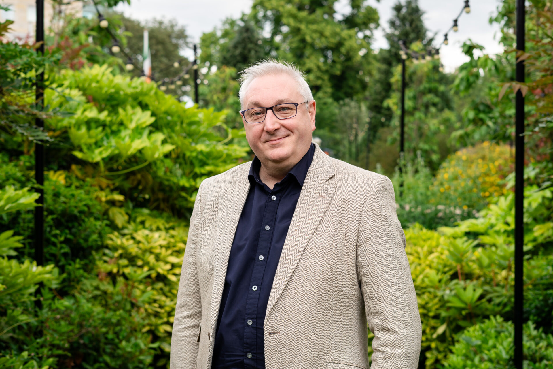 A man wearing spectacles, a light coloured jacket and a dark collared shirt