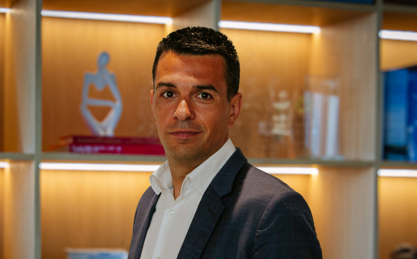 Picture of a man wearing a dark suit and white shirt with a glazed cabinet in the background