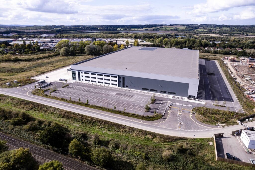Aerial view of a large modern industrial building surrounded by a wooded area and grassland