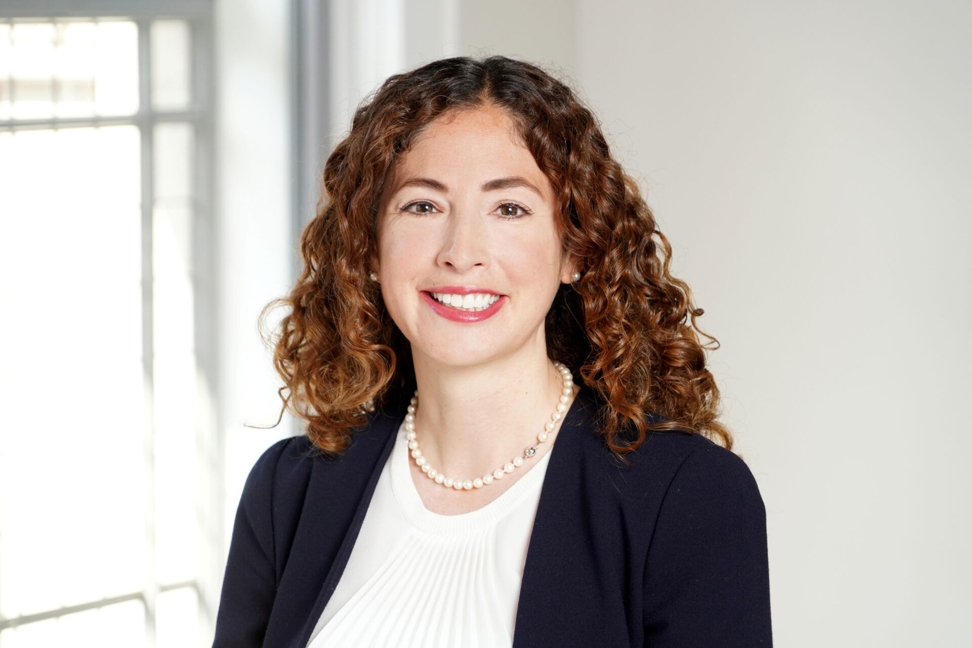 A smiling white woman with long curly brown hair and red lipstick wearing a white top, a blue jacket and a pearl necklace
