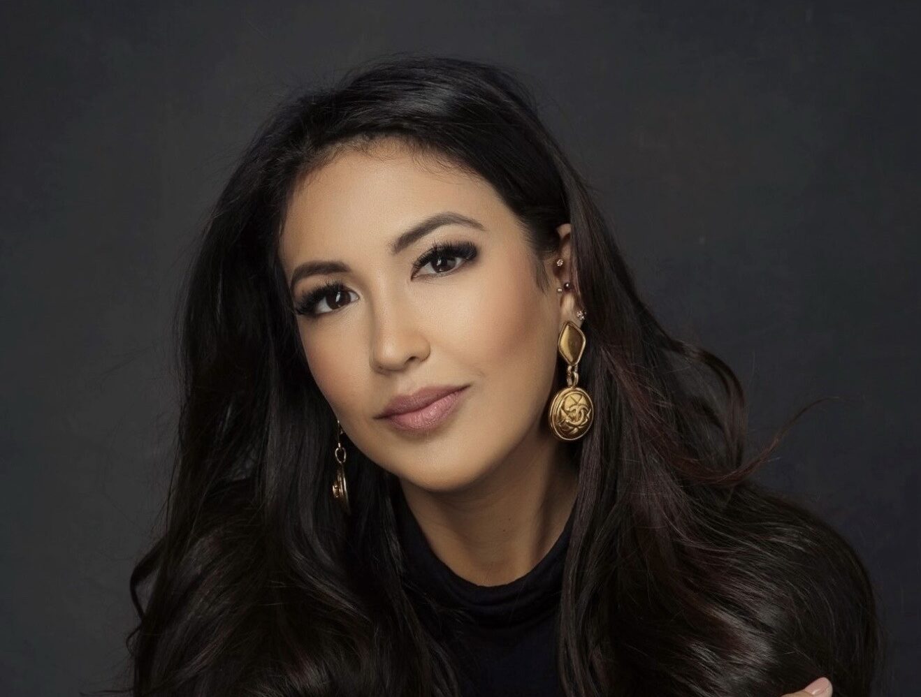 A woman with dark long hair wearing a dark top and gold earrings