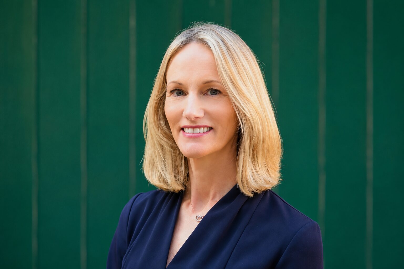 A woman with long blonde hair wearing a dark blue shirt in front of a green background
