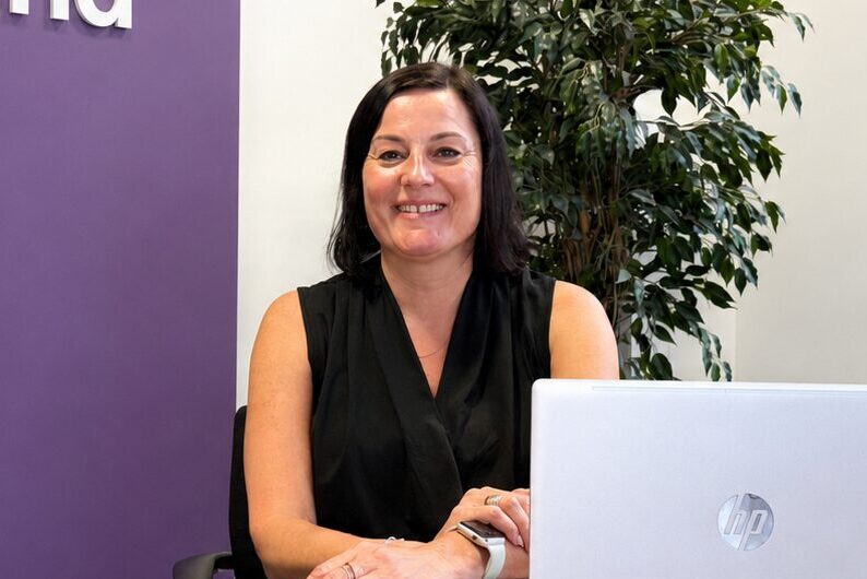 A woman with dark hair and a black shirt sat behind a desk with a computer in front of her