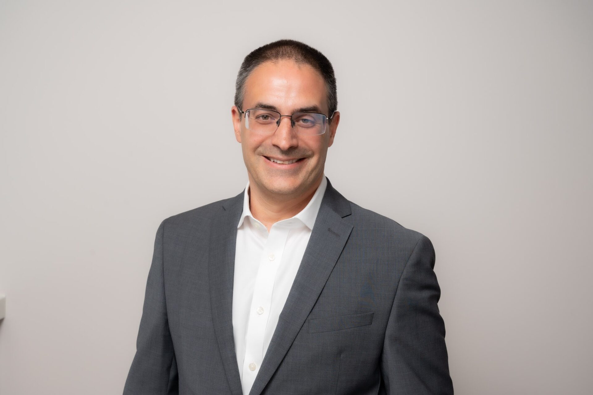 A smiling man with short hair wearing glasses, a grey suit jacket and a white collared shirt