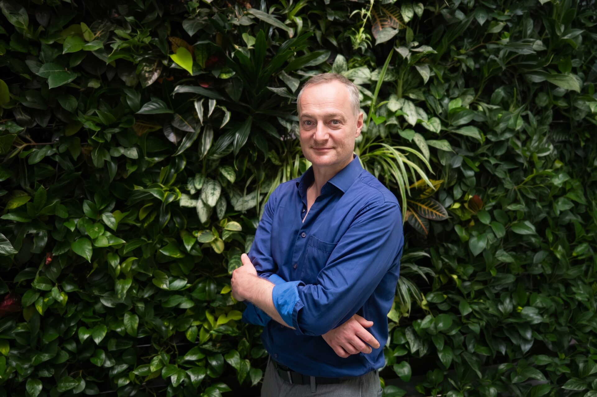 A smiling man wearing a blue shirt stood infront of a green bush