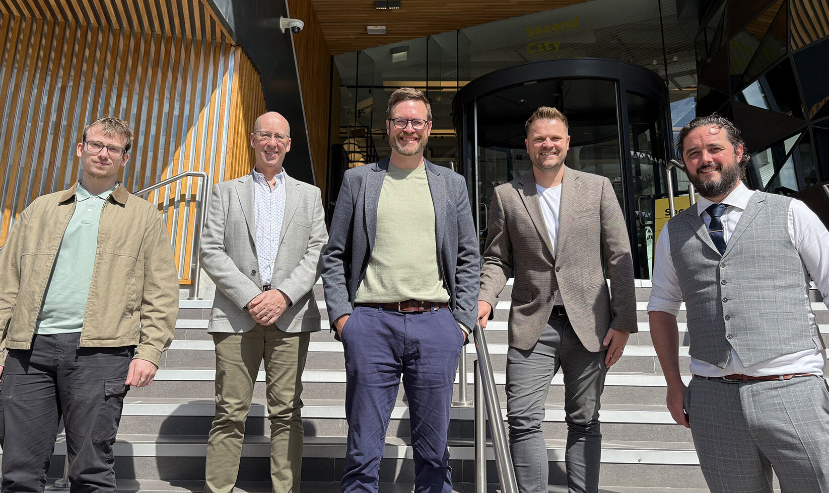 Four smiling men wearing smart clothing stood outside the entrance to a building