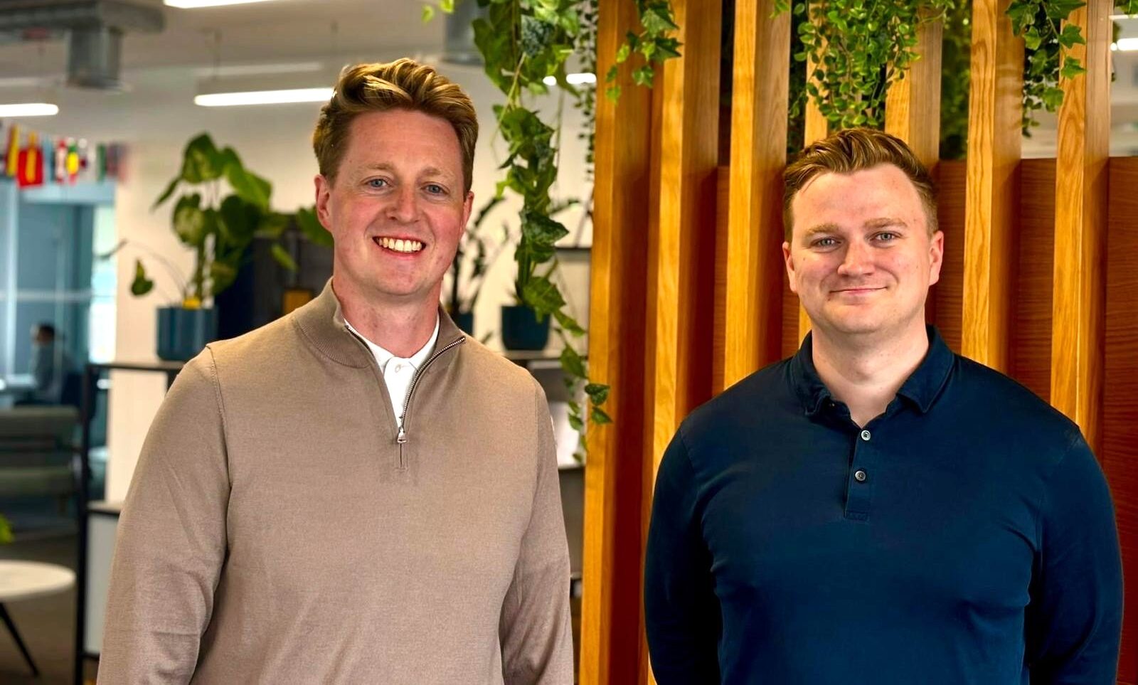 Two white males stood in front of a wooden background with foliage above their heads