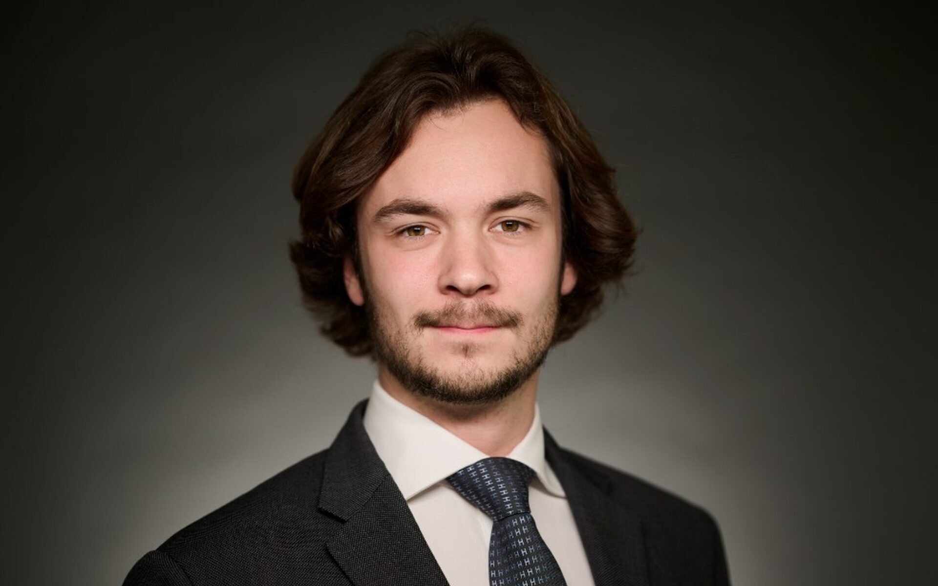 A white male with long brown hair and a beard wearing a dark suit jacket, a white shirt and a dark coloured tie