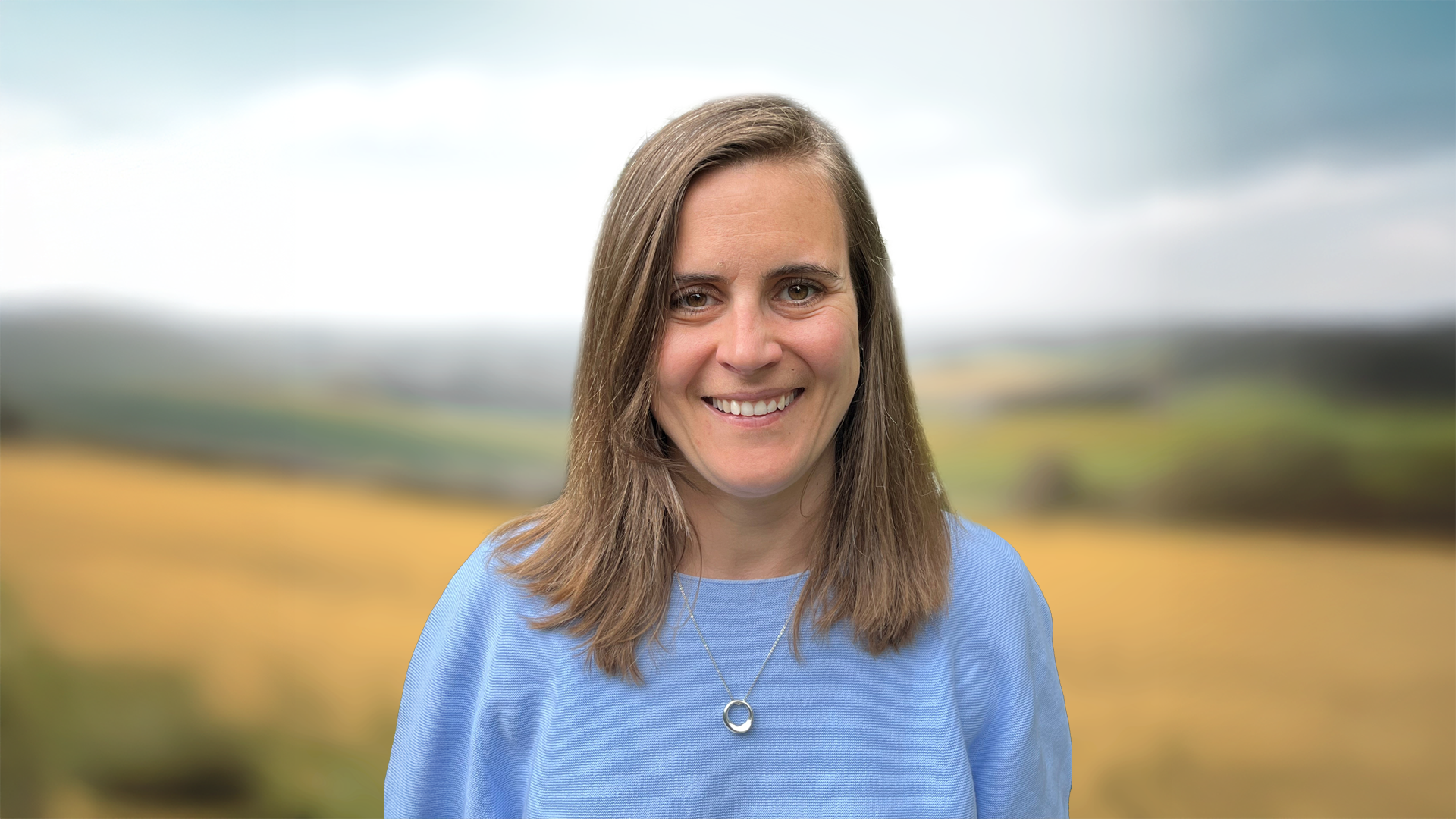 A woman with long brown hair wearing a blue shirt with a field in the background