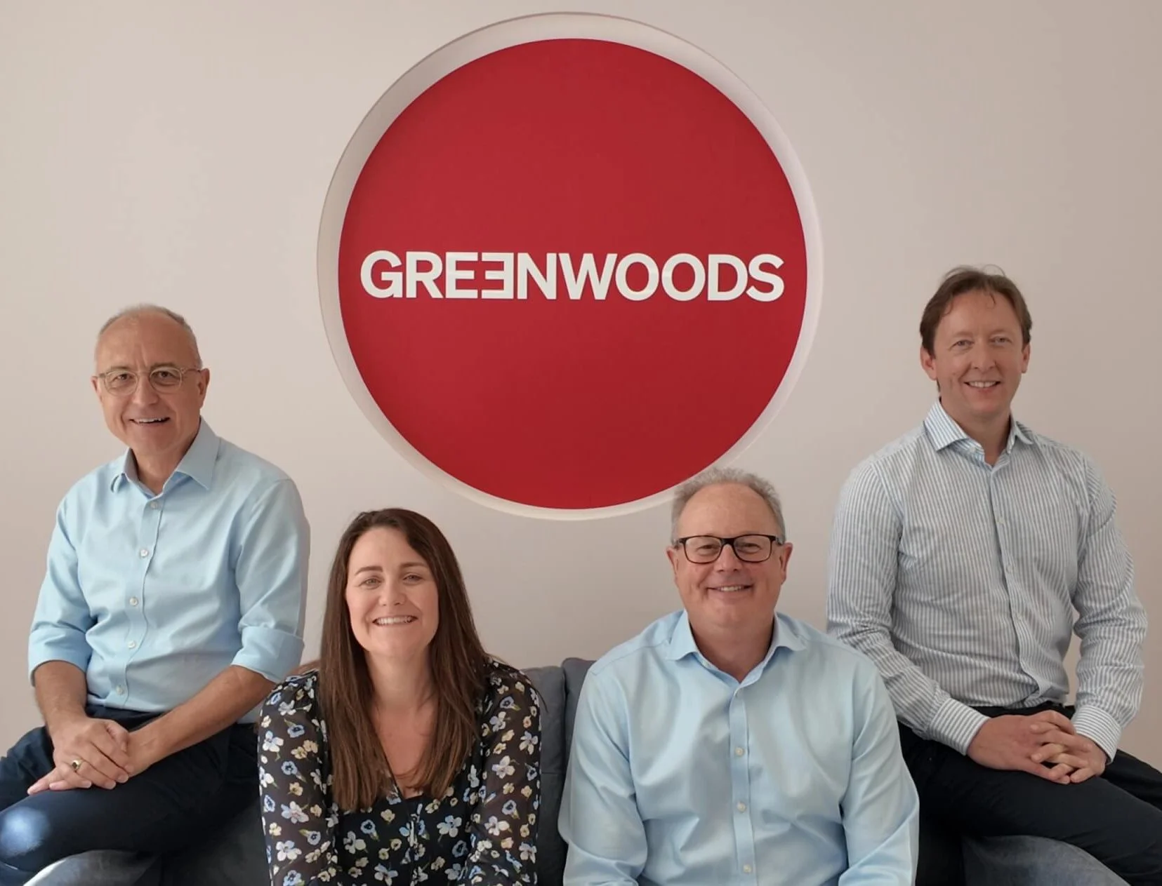 Three men and a women with a large red Greenwoods logo behind them