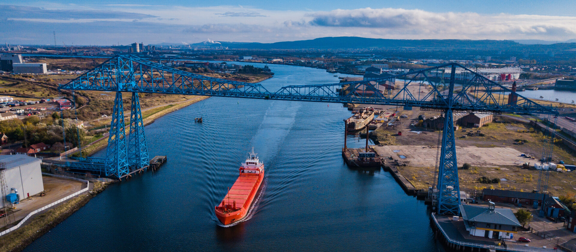 A red ship passing down a waterway under a large blue bridge