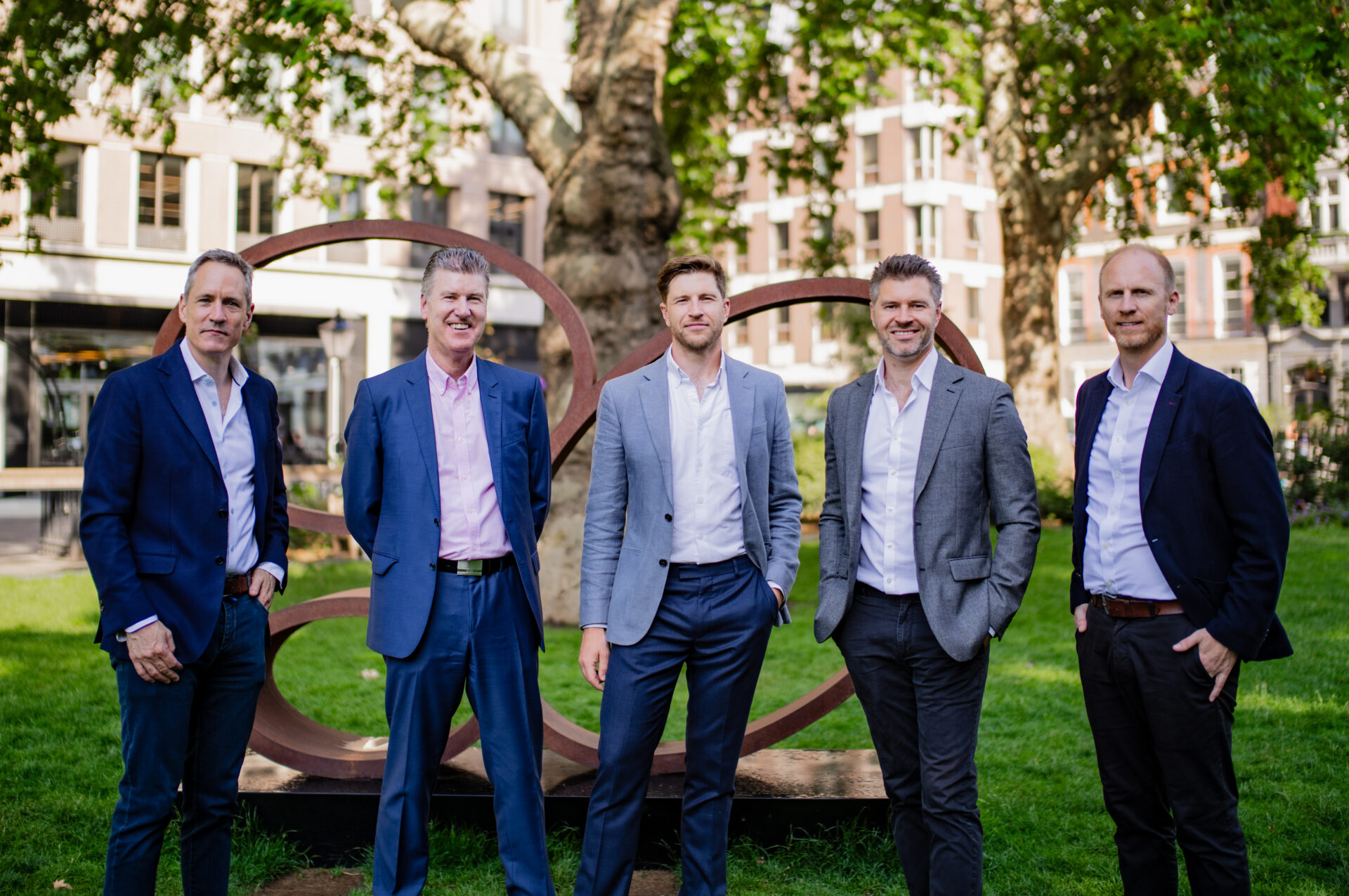 Five men wearing suits stood in a row in a park