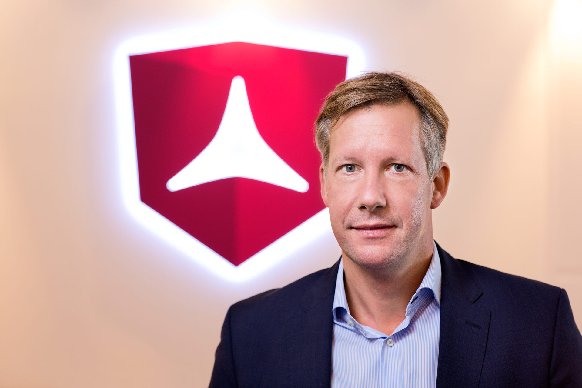 A man wearing a dark suit jacket and light coloured shirt in front of a red logo