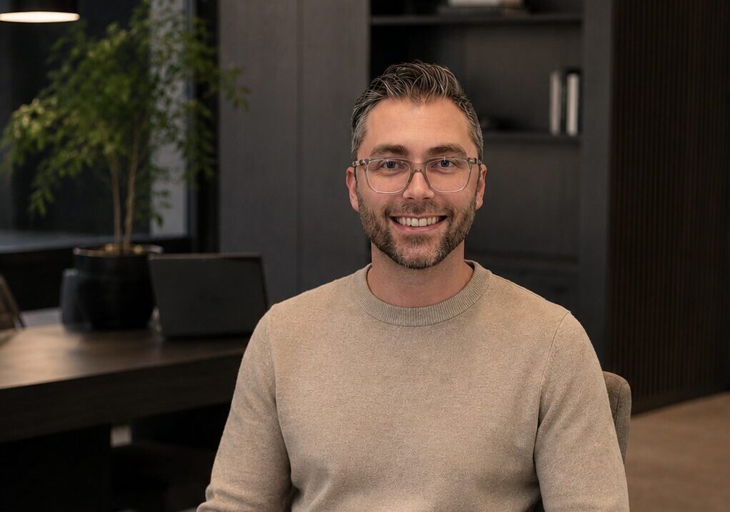 A smiling man with a beard wearing glasses sat in a chair