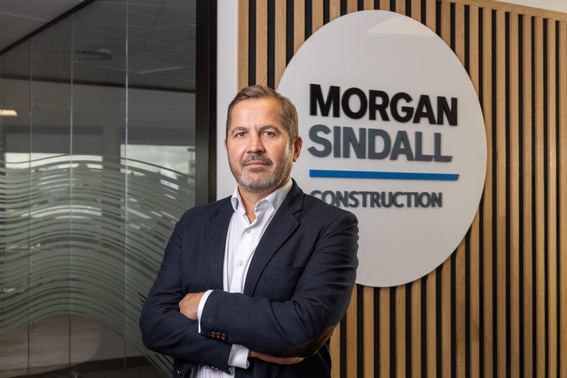 A man wearing a dark suit jacket with his arms crossed in front of a Morgan Sindall sign