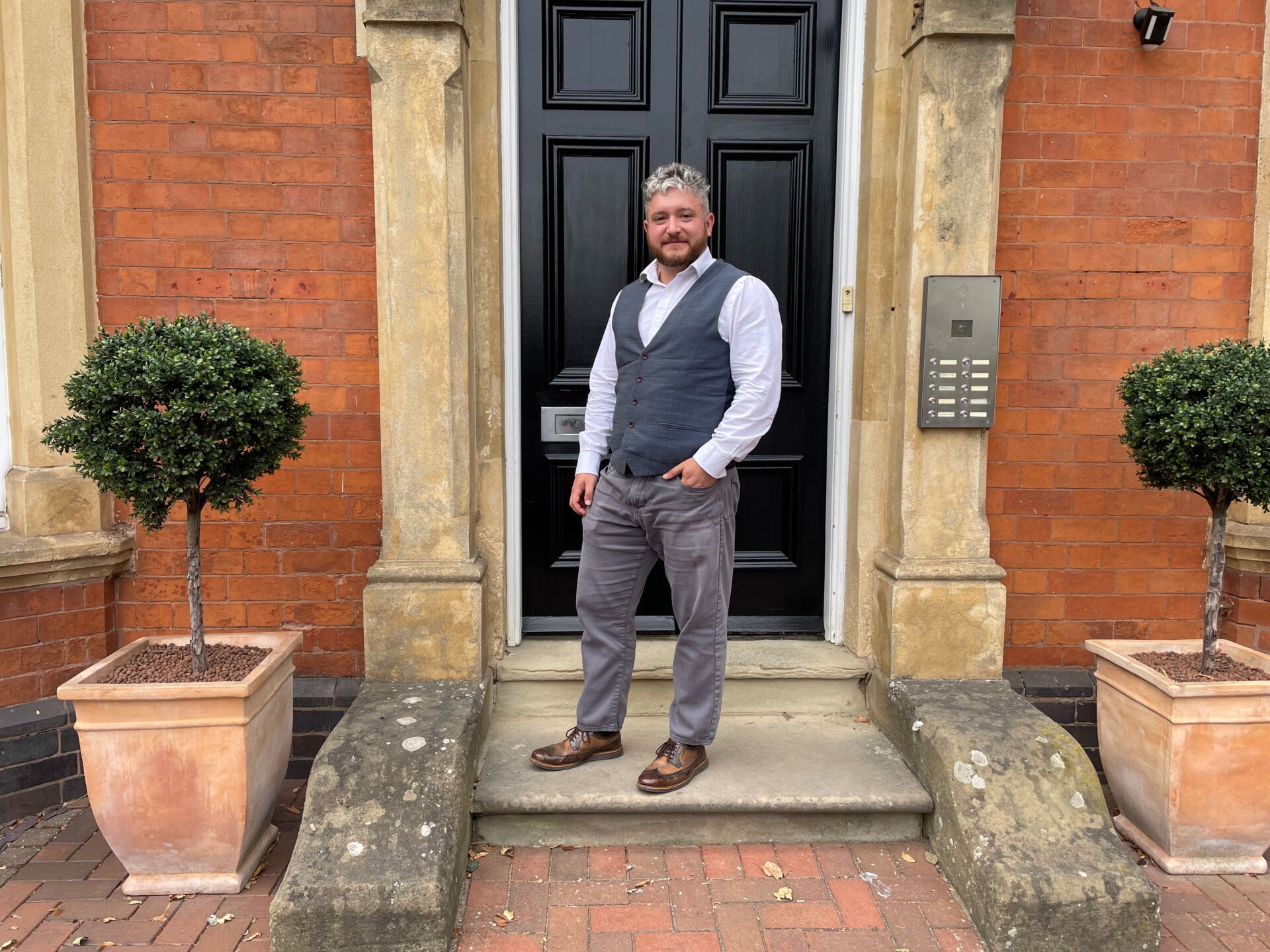 A man wearing a waist coat and white shirt stood in front of an ornate doorway
