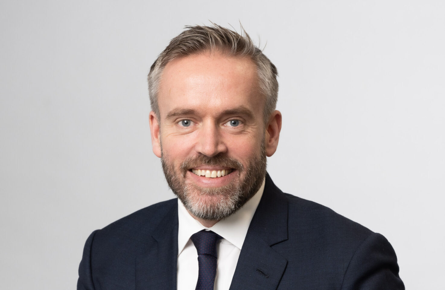 A smiling man with a beard wearing a dark suit jacket, a white shirt and a tie