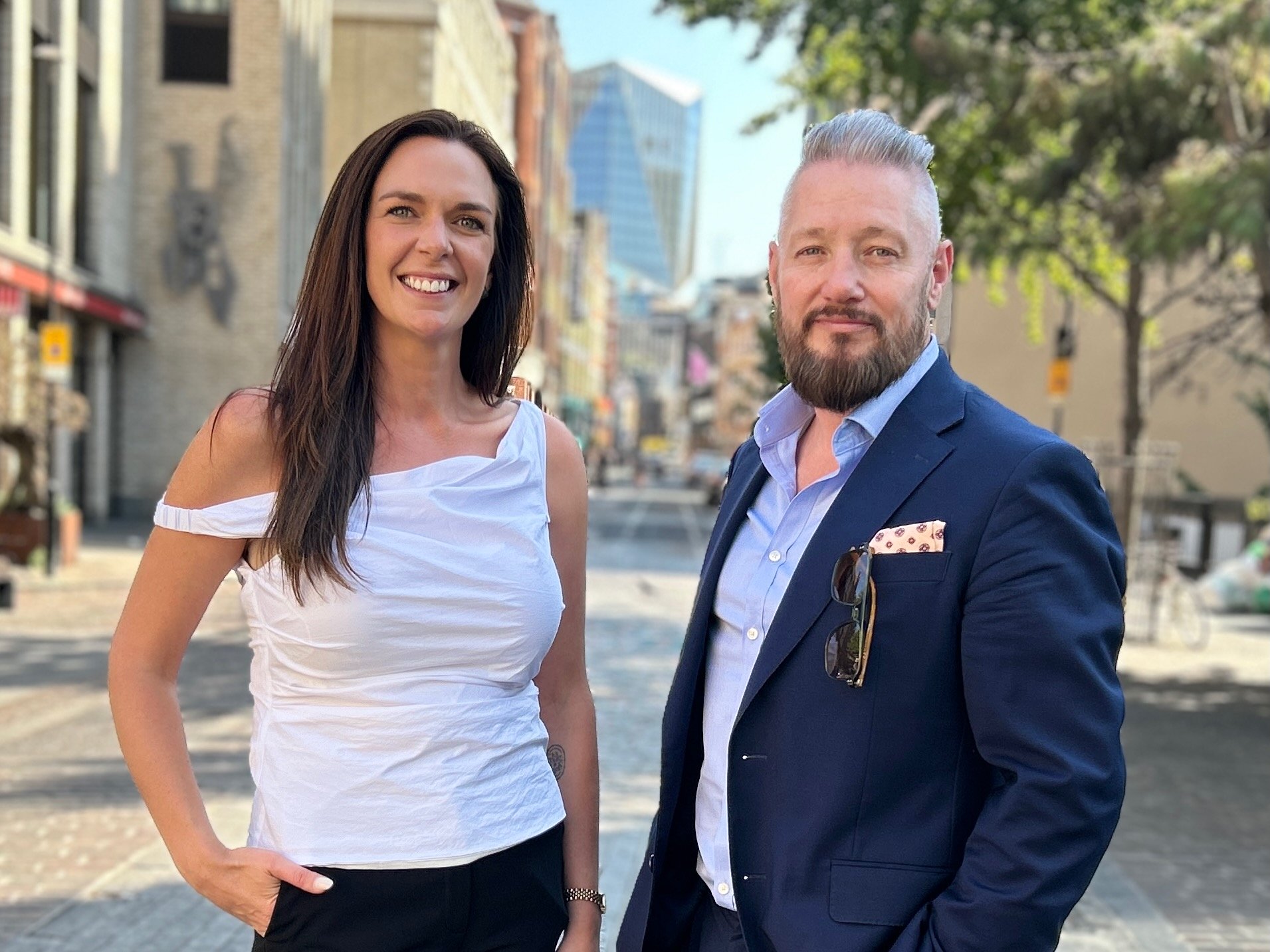 A woman wearing a white shirt next to a man with a beard wearing a dark suit jacket