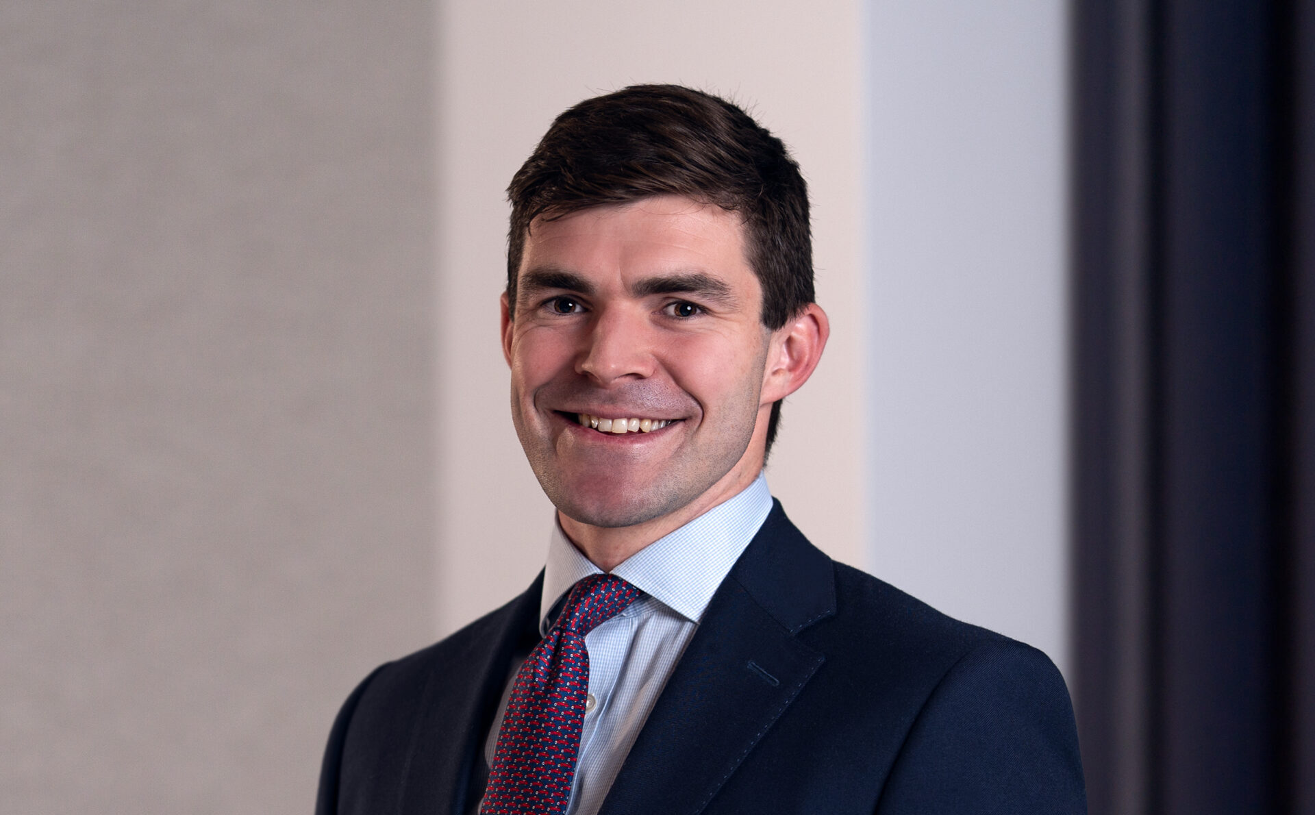 A smiling man with dark hair wearing a dark suit and a tie