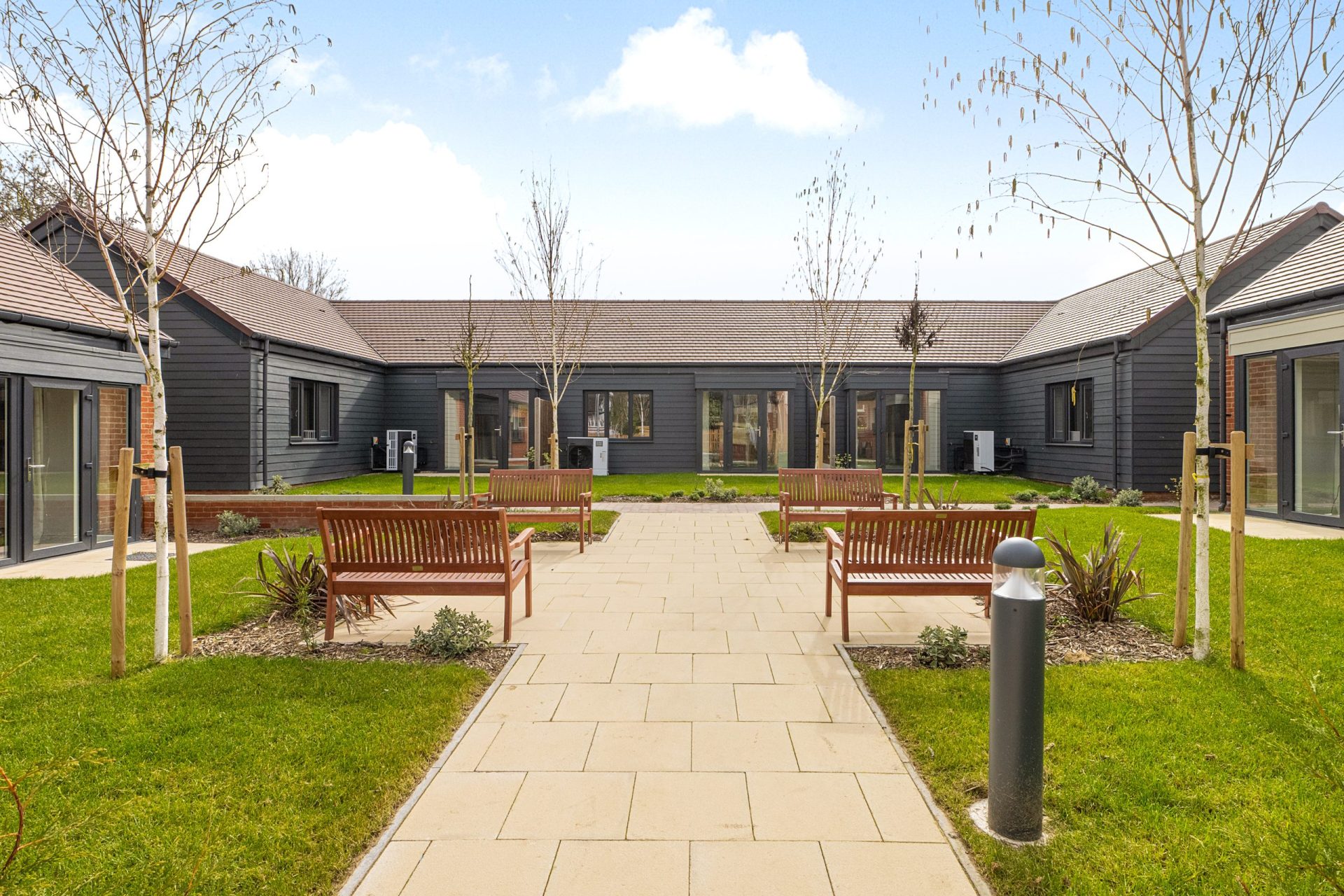 A connected series of grey wood panelled low rise buildings with a path and lawns in front