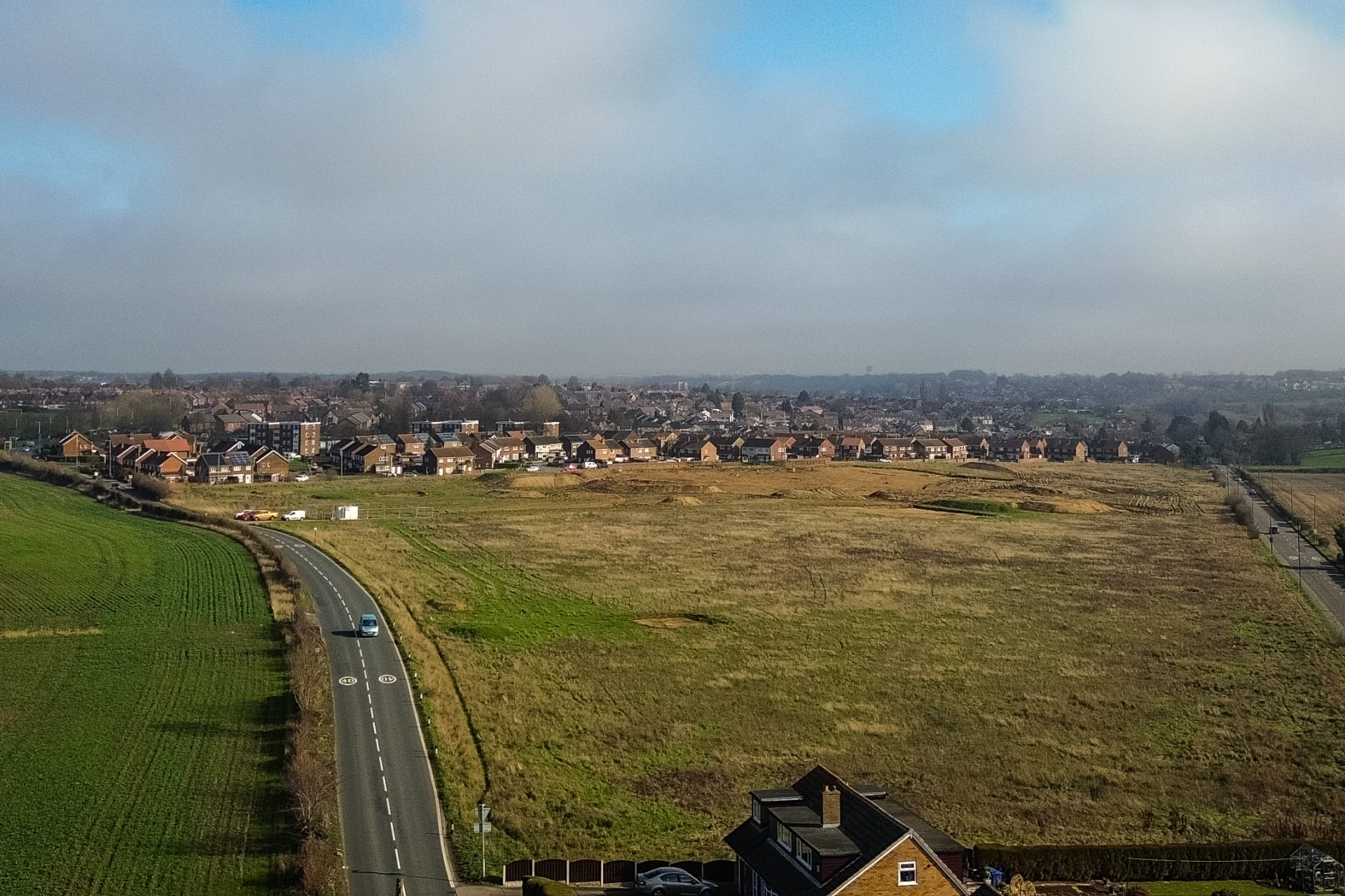Aerial view of a rural road beside open development land and a suburban housing estate