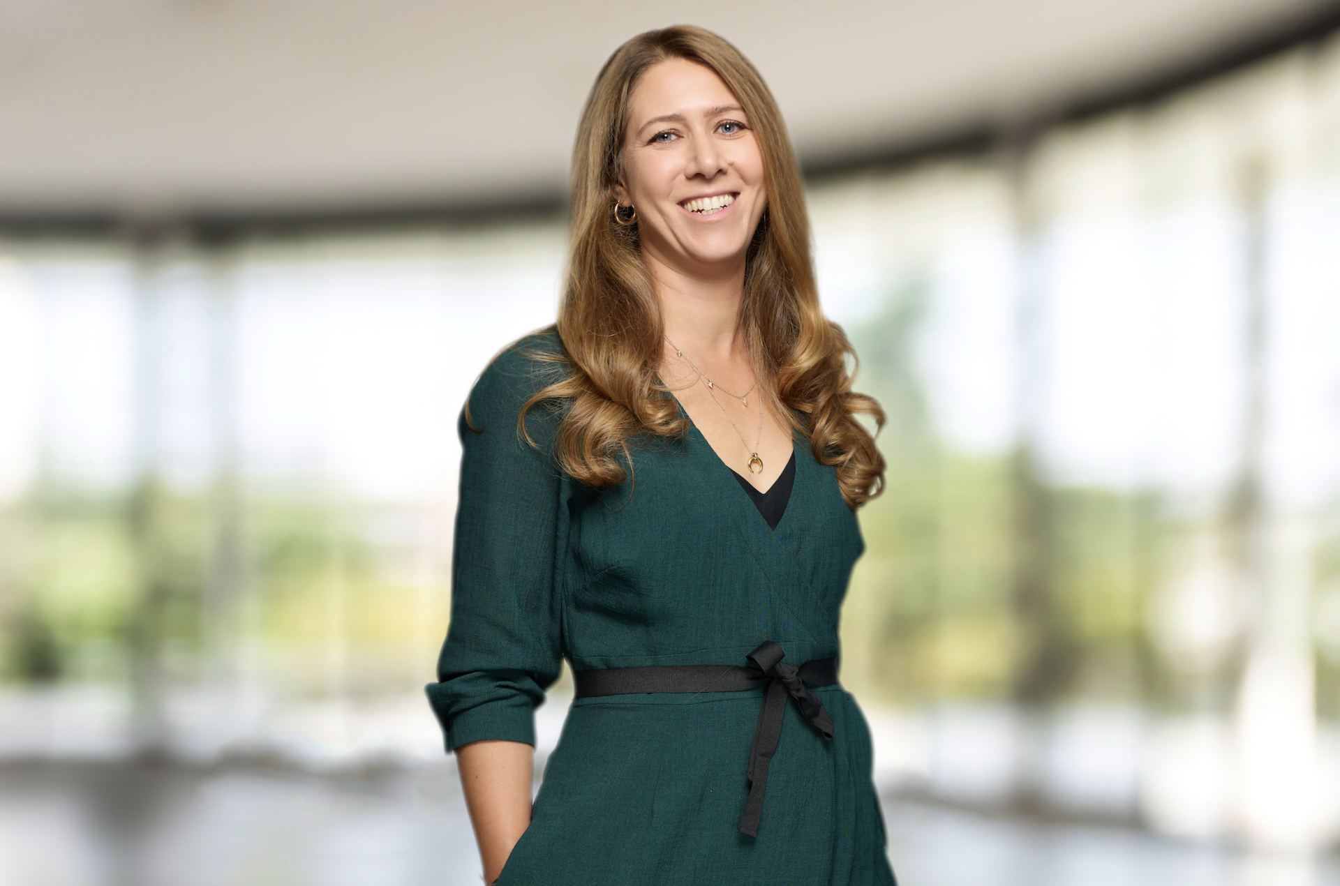 Alex Griffiths standing in a dark green dress against a blurred window background.