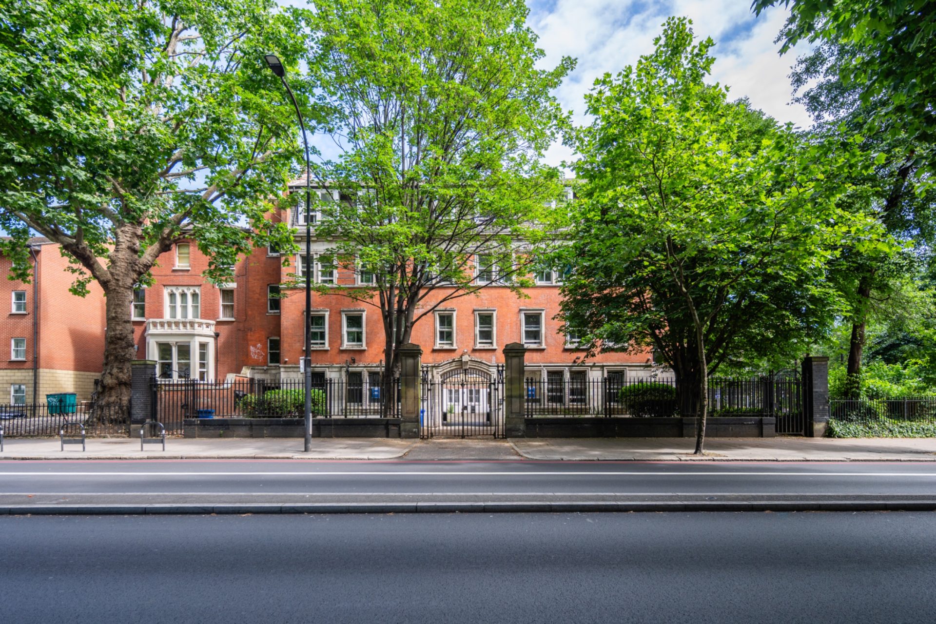 Tree-lined red-brick building behind black railings on an urban street