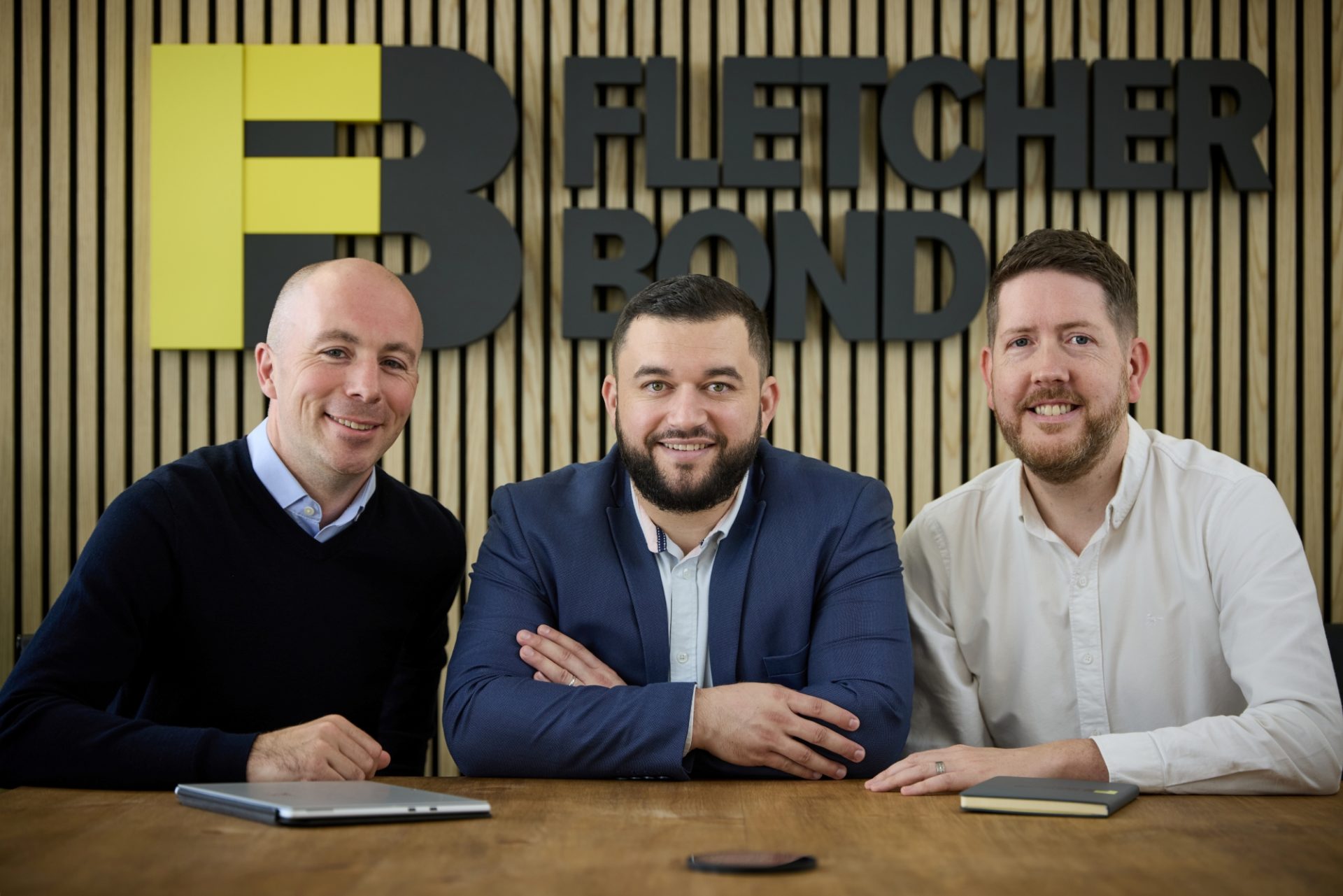 Three men sit at a table beneath a Fletcher Bond sign in the firm's Manchester office.