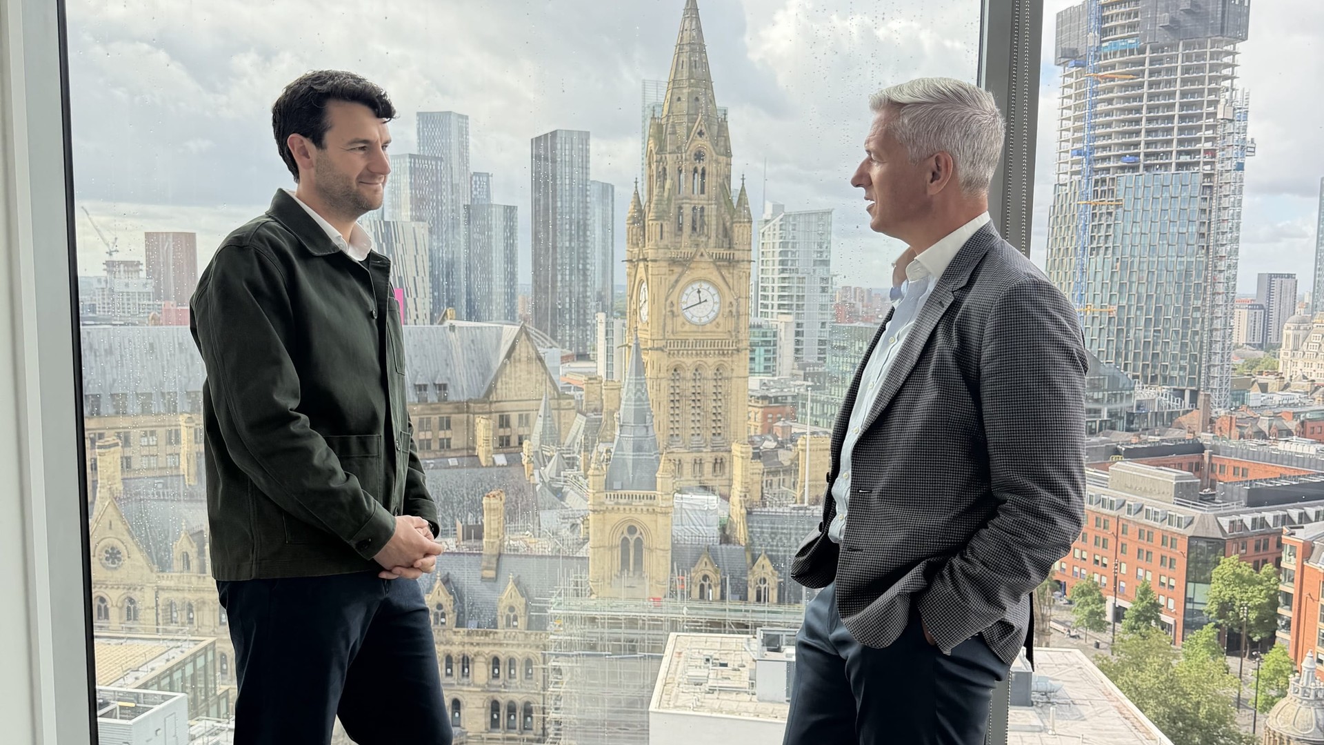 Two men stand by a window overlooking Manchester, with the Town Hall clock tower behind them.