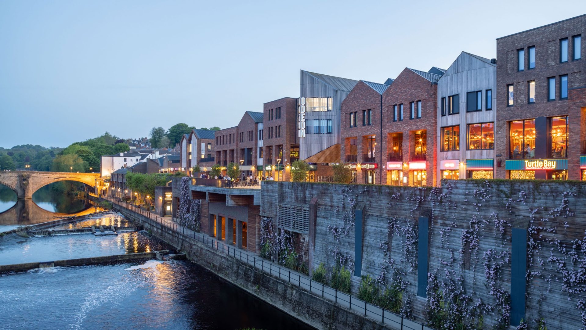 Modern riverside buildings and restaurants beside a weir and stone bridge at dusk