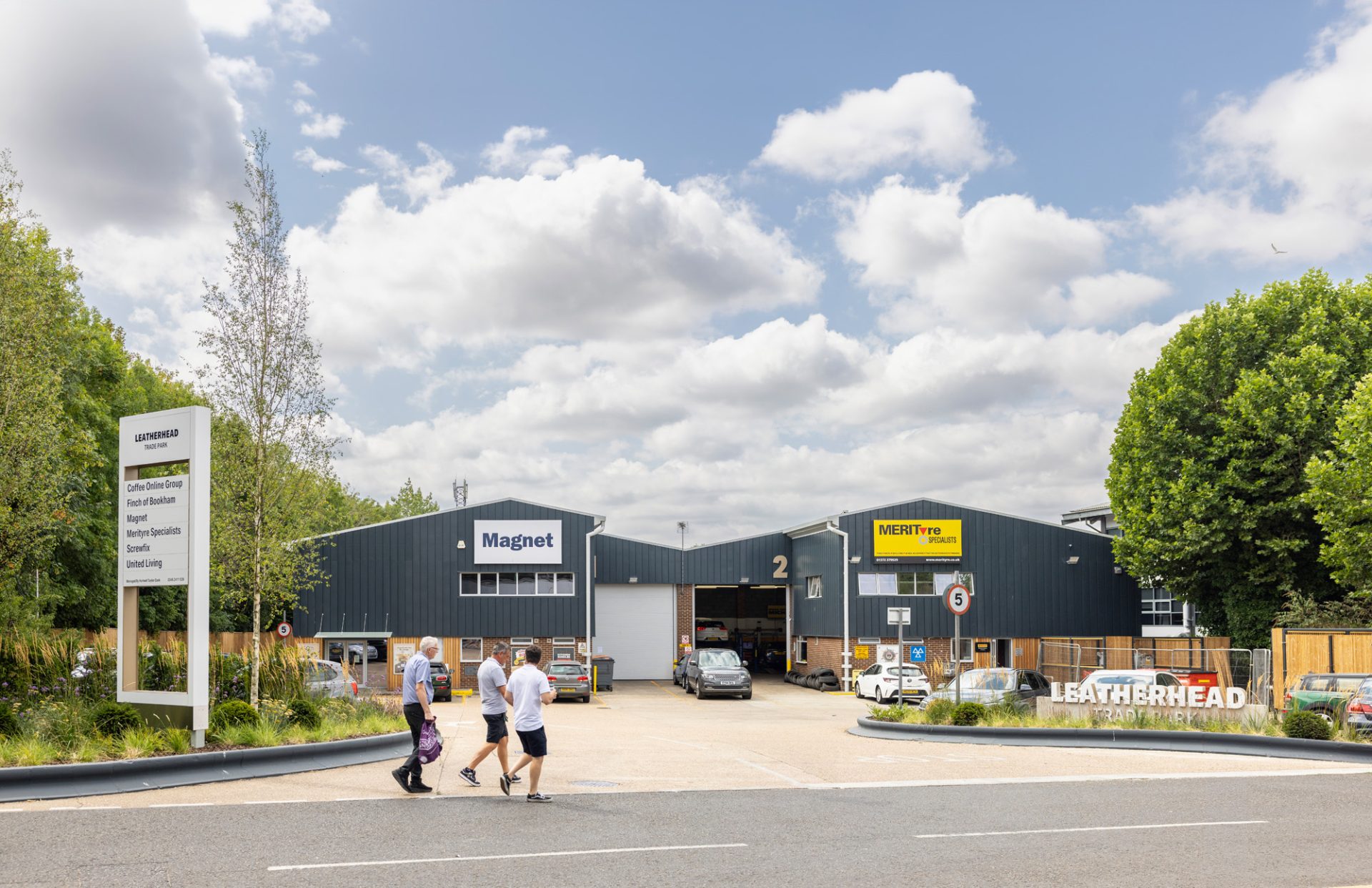 People walk past refurbished industrial units at a landscaped logistics estate under a cloudy sky